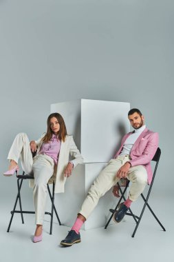 Gri, minimalist moda küplerinin yanındaki sandalyelerde oturan şık iş elbiseli genç çift.