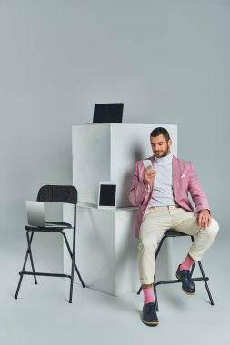 elegant man in lilac blazer sitting on chair with smartphone near devices on white cubes on grey clipart