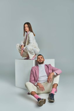 man in lilac blazer sitting on floor near woman in elegant suit posing on white cube on grey clipart