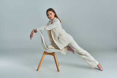 full length of expressive woman in white suit posing with armchair and looking at camera on grey clipart