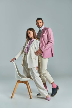 Pastel resmi giyinen genç ve zarif bir çift gri koltukta poz veriyor, modern iş tarzı.