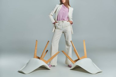 cropped view of woman in white suit standing with hands in pockets near overturned armchairs on grey clipart