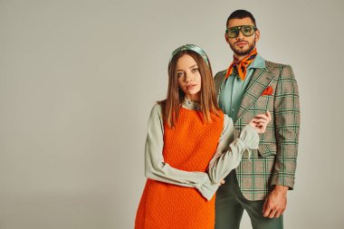 Turuncu elbiseli ve ekose ceketli genç ve şık bir çift gri, eski moda kameralara bakıyor.