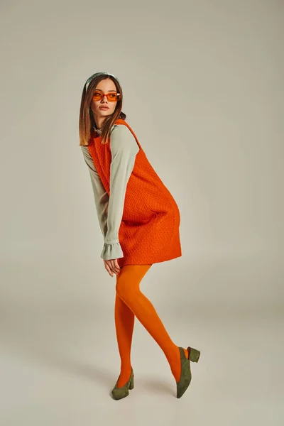 full length of vintage style woman in orange dress, tights and sunglasses looking away on grey