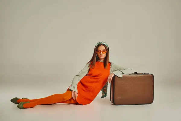 young model in fashionable vintage attire and sunglasses sitting near vintage suitcase on grey