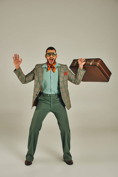 весёлый мужчина в клетчатой куртке и солнечных очках с винтажным чемоданом на сером, ретро-вдохновлённом образе жизни