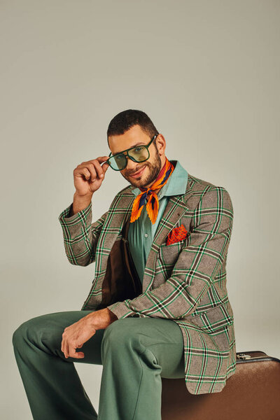 счастливый человек в ретро-одежде сидит на винтажном чемодане и улыбается в камеру на сером фоне