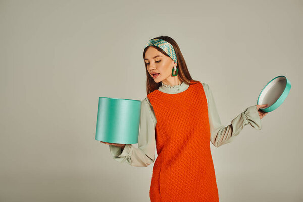 Любопытная женщина в оранжевом платье и ободок глядя в подарочной коробке на сером, ретро моды фотографии