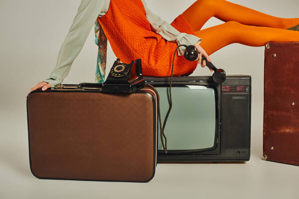 cropped view of woman in orange dress sitting on vintage suitcase and tv set on grey, retro style