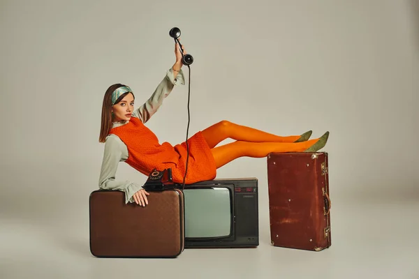 穿着时髦复古服装的年轻女人 坐在电视机上的老式电话和灰色行李箱上 — 图库照片
