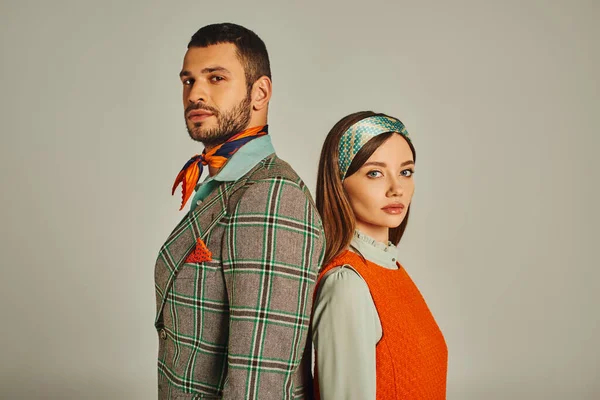 young couple in stylish old-fashioned attire standing back to back and looking at camera on grey
