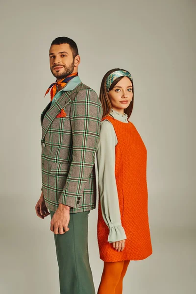 positive and trendy couple in orange dress and plaid jacket looking at camera on grey, retro fashion