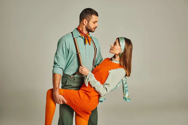 trendy man in suspenders flirting with woman in orange dress on grey, retro-inspired fashion