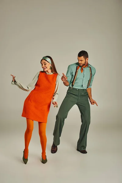 full length of young trendy couple in retro attire dancing on grey, retro vibes and entertainment