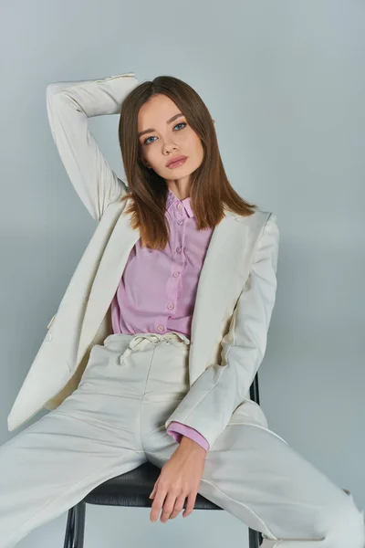 modern woman in white suit sitting on chair with hand behind head and looking at camera on grey