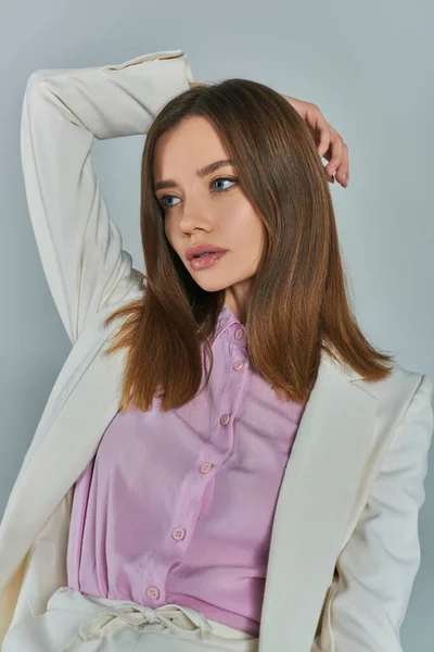 stock image young charming woman in white blazer looking away with hand behind head on grey, fashion and style