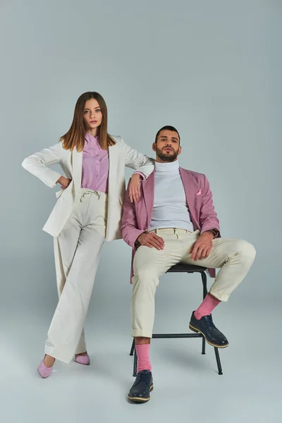 stock image woman in white formal suit posing with hand on hip near man in lilac blazer sitting on chair on grey