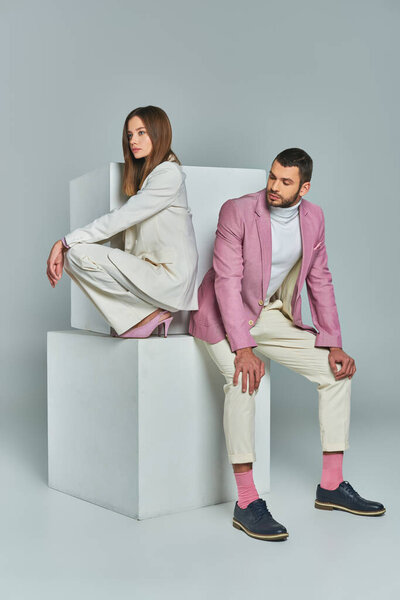 full length of young fashionable couple in elegant formal attire posing on white cubes on grey