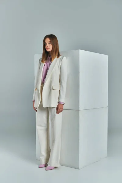 trendy business attire, modern woman in white elegant suit standing near cubes on grey, full length