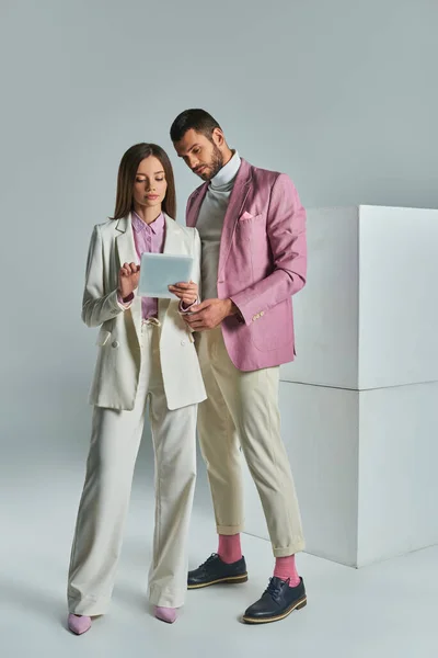young business couple in stylish formal attire looking at digital tablet near white cubes on grey