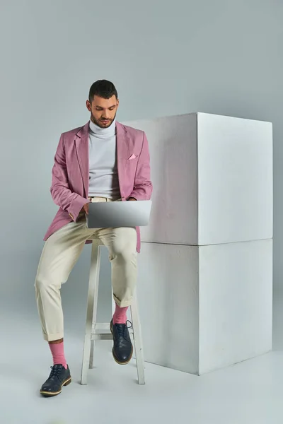 穿着丁香夹克的年轻商人坐在凳子上 在灰色的白色立方体旁边的笔记本电脑上工作 — 图库照片