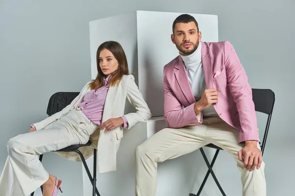 young couple in trendy formal wear sitting on chairs near cubes on grey, modern business fashion