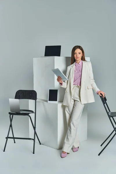 young woman in white suit holding laptop near chairs and digital devices on white cubes on grey