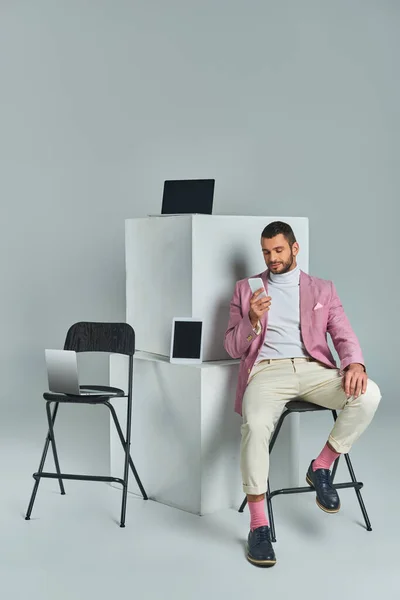 stock image elegant man in lilac blazer sitting on chair with smartphone near devices on white cubes on grey