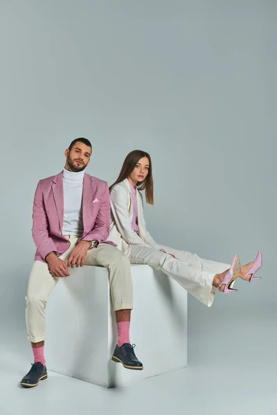 young business couple in elegant formal wear sitting on white cube and looking at camera on grey