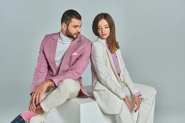 stock image young and fashionable couple in elegant suits sitting on white cube on grey backdrop in studio