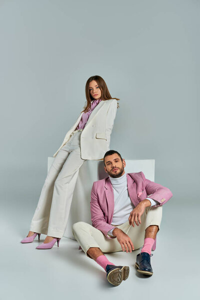 man in lilac blazer sitting on floor near woman in stylish suit posing near white cube on grey
