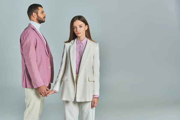 stock image young woman in white suit holding hands with man in lilac blazer and looking at camera on grey