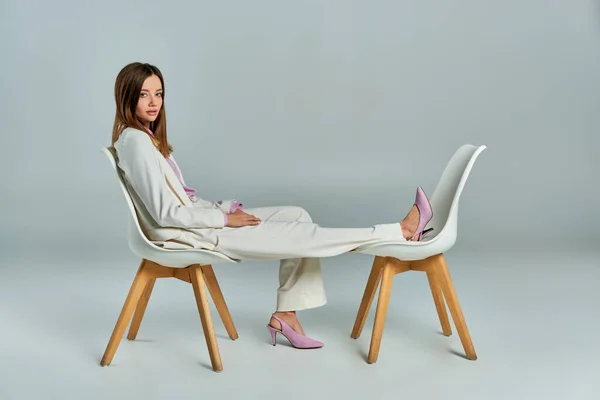 stock image young and elegant woman in white suit posing on armchairs on grey, minimalistic business fashion