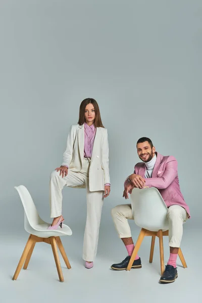 stock image joyful man in lilac blazer sitting and looking at camera near woman posing with armchair on grey