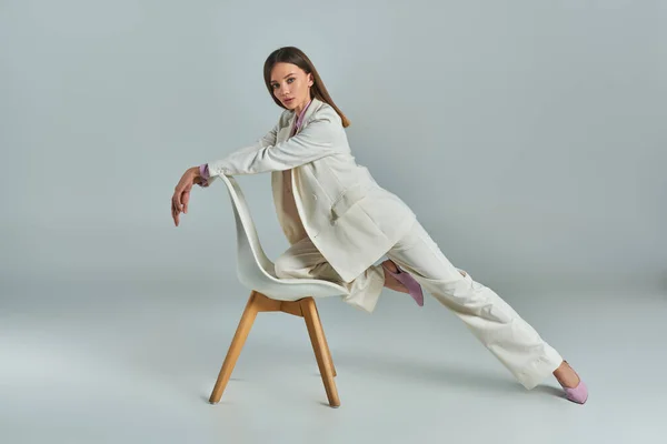 stock image full length of expressive woman in white suit posing with armchair and looking at camera on grey