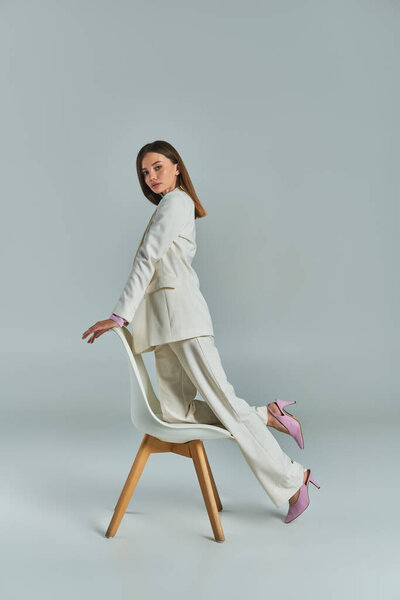 modern woman in white elegant suit looking at camera and posing with armchair on grey, full length