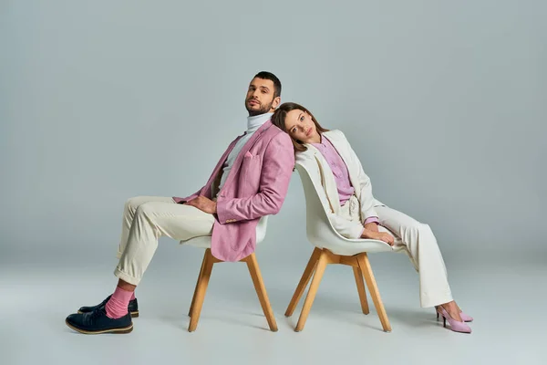 young couple in stylish formal wear sitting back to back in armchairs and looking at camera on grey