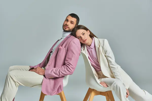 stock image elegant couple in formal wear sitting in armchairs and looking at camera on grey, modern fashion