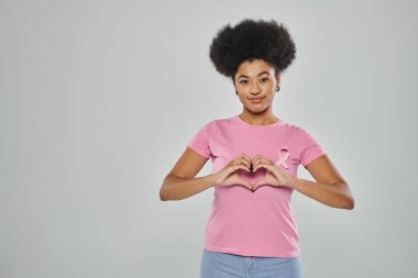 Göğüs kanseri farkındalığı, gri arka planda pembe kurdeleli Afro-Amerikan kadın, kalp işareti, gülümseme