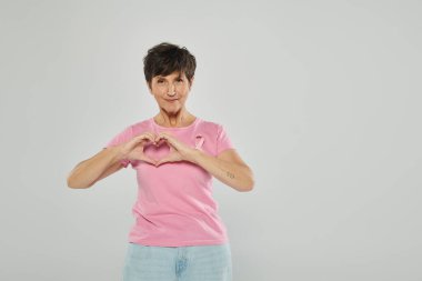 Göğüs kanseri farkındalığı, pembe kurdeleli mutlu olgun kadın, gri perde arkası, portre, kalp işareti
