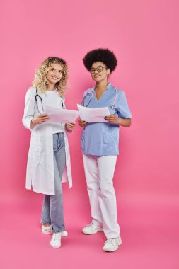 cheerful female oncologists, interracial doctors on pink backdrop, breast cancer awareness clipart