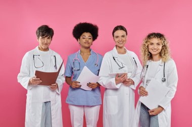 Sırıtkan çok ırklı doktorlar ellerinde kağıt klasörler ve pembe ve göğüs kanserine izole edilmiş dijital tablet tutuyorlar.