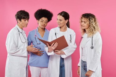 Sırıtkan doktor elinde pembe ve meme kanserine izole edilmiş çok ırklı meslektaşlarının yanında kağıt klasör tutuyor.