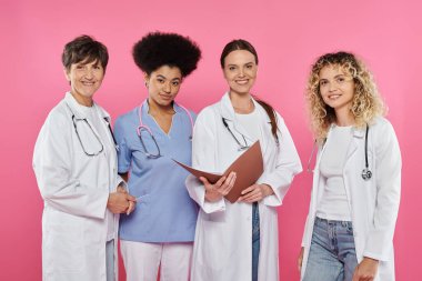 happy multiethnic doctors holding paper folder and looking at camera isolated on pink, breast cancer clipart