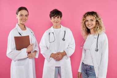 smiling female doctors in white coats looking at camera isolated on pink, breast cancer awareness clipart