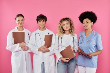 happy interracial oncologists with paper folders looking at camera isolated on pink, breast cancer clipart