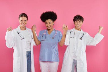 excited multiethnic doctors oncologists holding hands isolated on pink, breast cancer month clipart