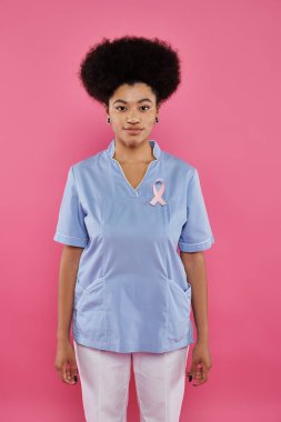 Göğüs kanseri kurdelesi takmış Afro-Amerikan kadın doktor pembe üzerinde tek başına duruyor.