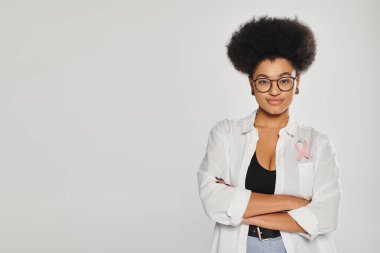 Göğüs kanseri pembe kurdeleli Afro-Amerikalı bir kadın. Gri renkte izole bir şekilde kollarını kavuşturuyor.