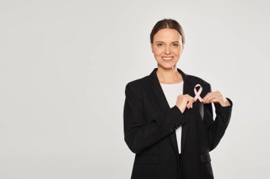 Takım elbiseli pozitif iş kadını, gri alanda izole edilmiş pembe göğüs kanseri kurdelesi taşıyor.